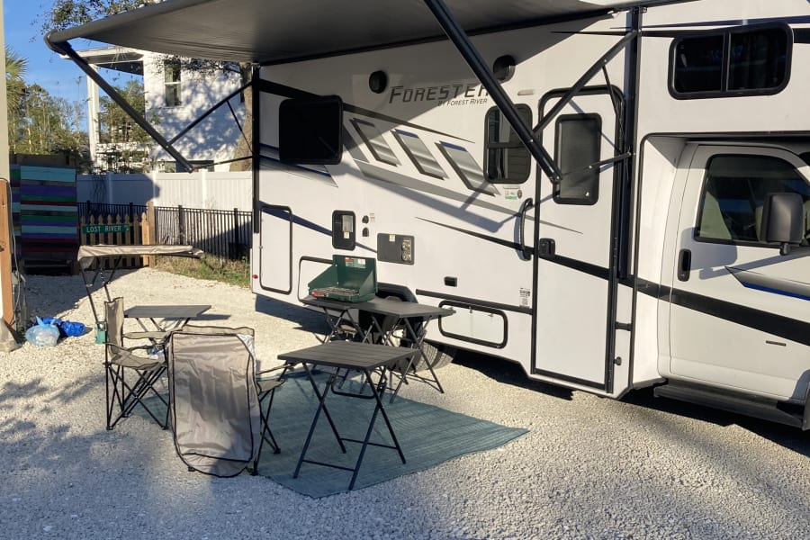 RV rental parked on gravel, with its awning extended over a comfy outdoor setup including a portable grill and folding chairs.