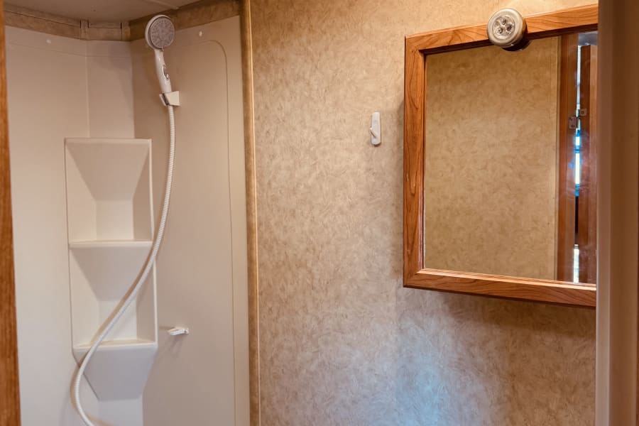 Inside an RV rental bathroom, featuring a compact tub/shower with handheld showerhead, vanity with sink & mirror, and overhead lighting.