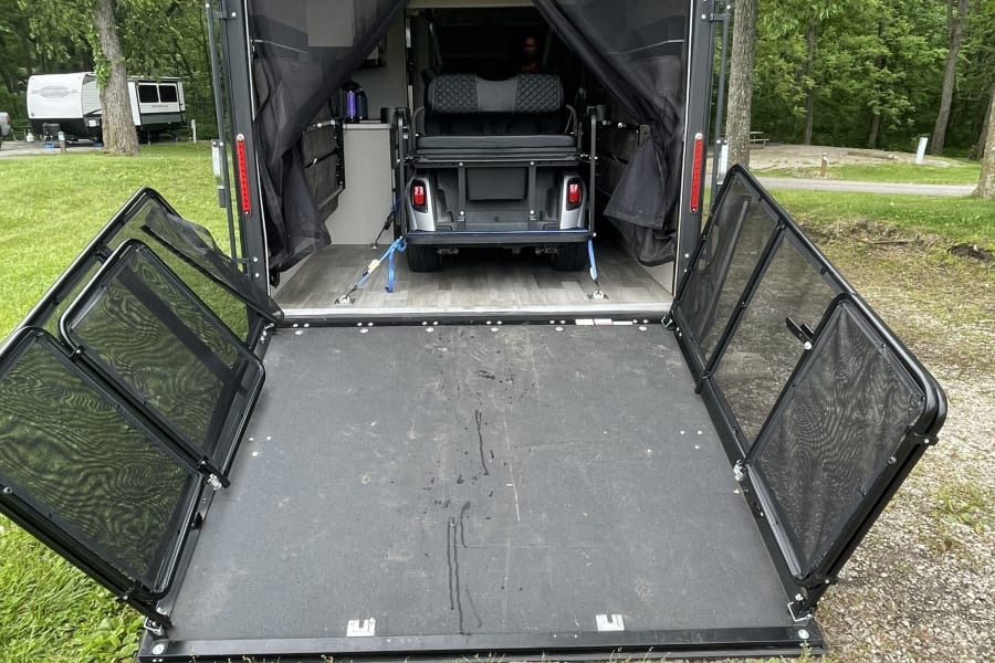 Toy hauler RV rental: ramp down as a patio, a golf cart secured inside at a wooded campground.