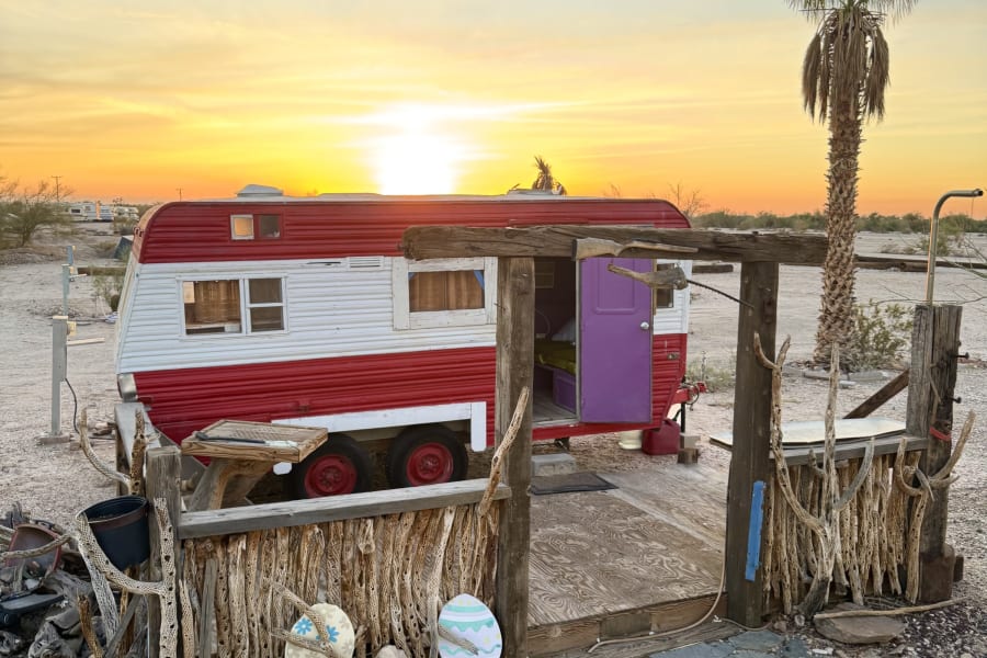 Vintage red & white rv rental set in the desert. Features a unique wooden porch, purple door, and a vibrant sunset.