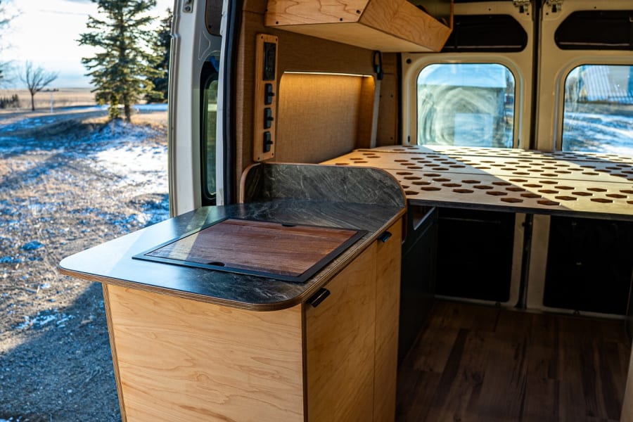 RV rental: Inside a modern wood campervan with kitchen, bed, and an open door revealing a snowy landscape.