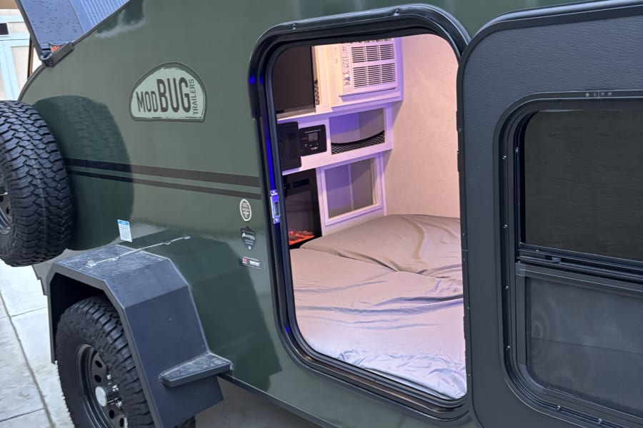 This dark green Modbug teardrop RV rental features an open door revealing a cozy bed, AC unit, and controls inside.