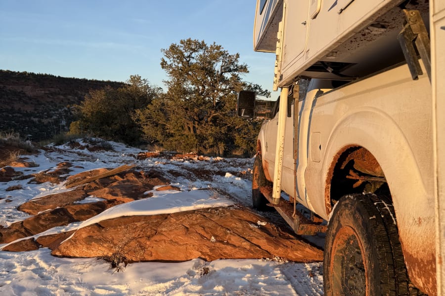 White RV rental, mud-splattered, parked on snow-dusted red rocks with a distant scenic view and blue sky at golden hour.