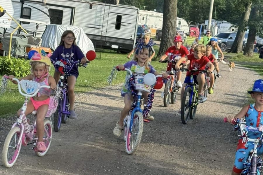 Children on bikes, decorated with red, white, & blue, parade past RV rental campers. Family fun at the campground.