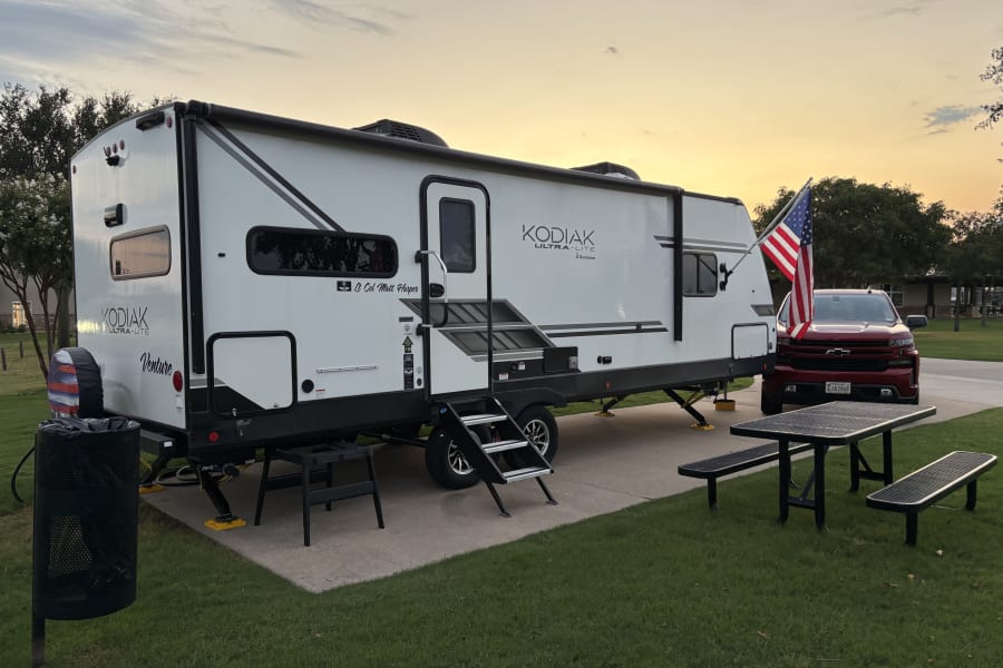 Kodiak travel trailer (possibly RV rental) at a campsite with a red truck, American flag, and picnic area.