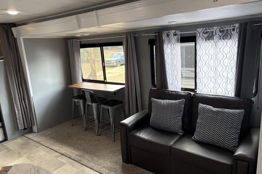 Cozy RV rental interior with black leather sofa, chevron pillows, bar table, and windows with grey & sheer white curtains.