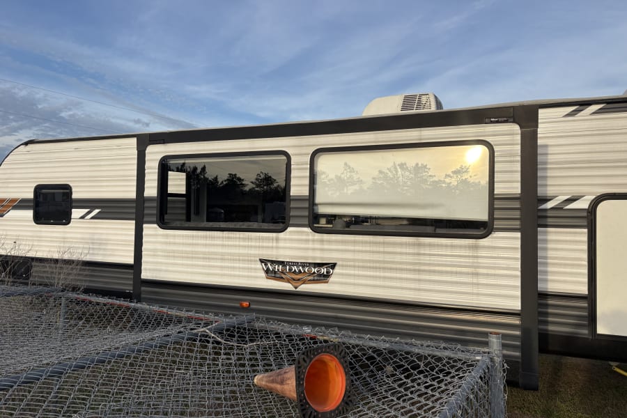 Wildwood travel trailer (rv rental) parked behind a chain-link fence with an orange cone. Sunny skies overhead.