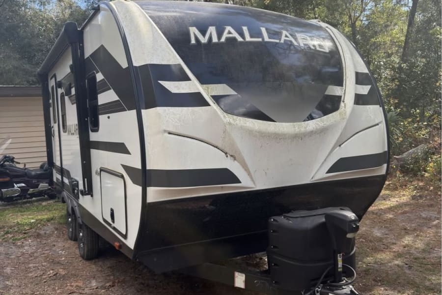 White and black Mallard travel trailer for rv rental, parked outdoors on dirt under trees, ready for your next adventure.