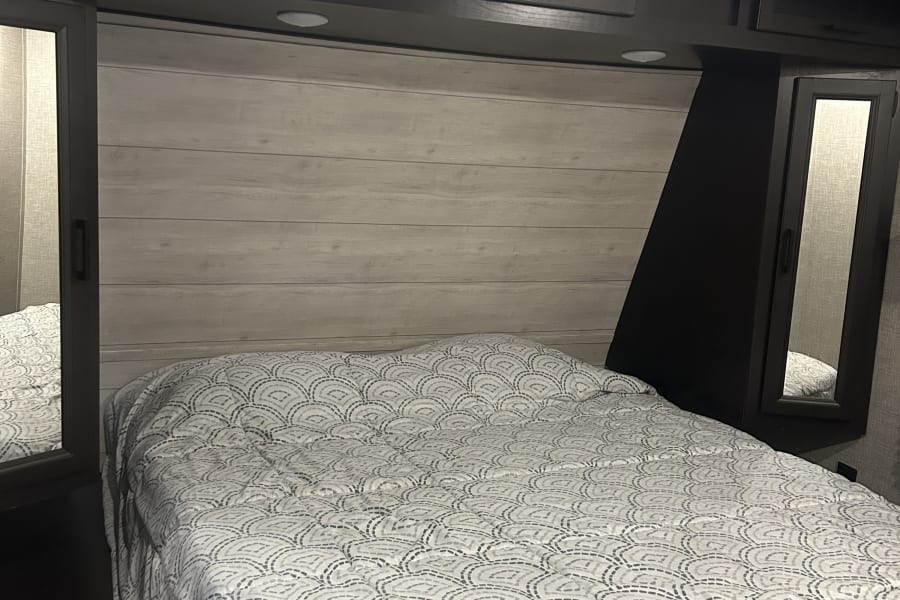 Experience a comfortable rv rental bedroom. Features include a patterned bedspread, wood-look headboard, and overhead cabinets.