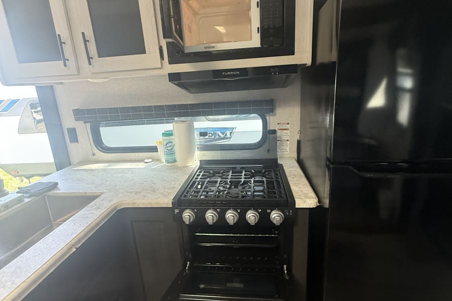 RV rental kitchen featuring an open microwave, stove, oven, sink, and black refrigerator. Light wood cabinets complete the space.