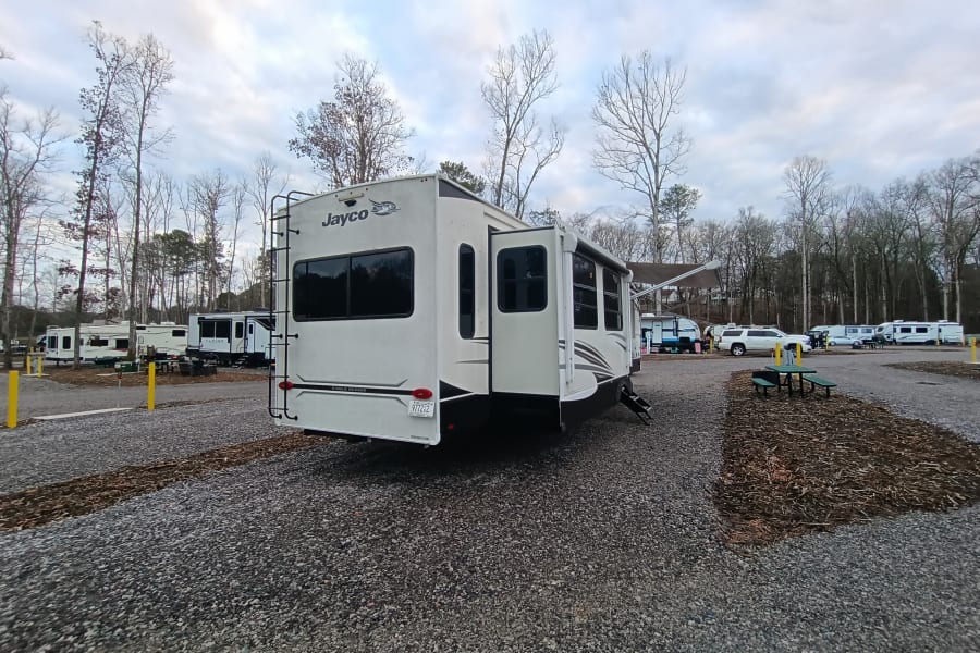 Spacious Jayco fifth-wheel RV rental, with slides out, parked on a gravel campsite among bare trees under a cloudy sky.