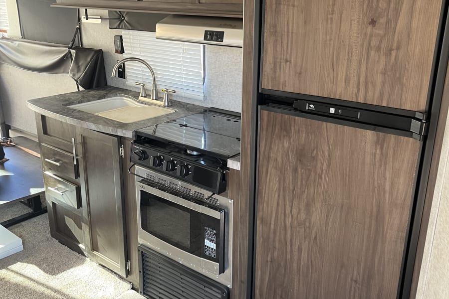 Modern RV rental kitchen features wood-look cabinets, sink, stove, microwave, fridge, and carpeted floor.