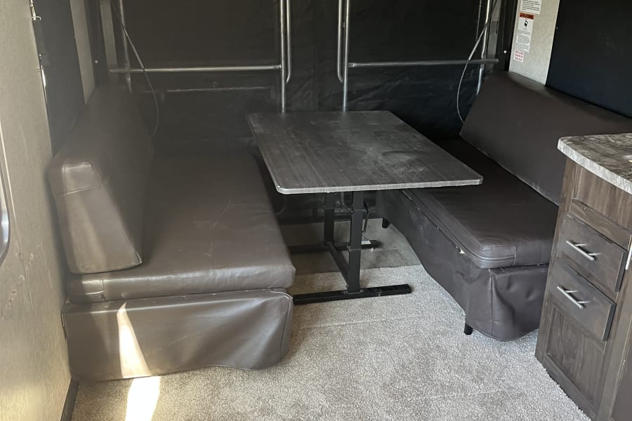 Interior of an RV rental showing a dinette with dark brown benches, a wood-look table, and light gray carpet. Includes toy hauler partition.