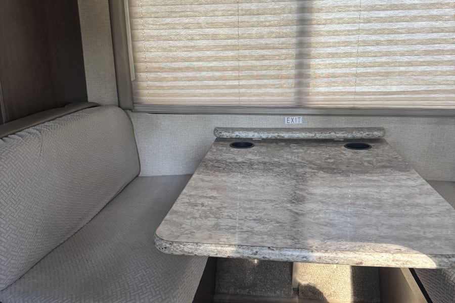 Inside an RV rental: a bright dinette area with patterned bench seating, a marble-look table with cup holders, and a window.