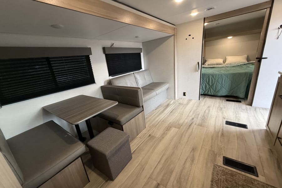 Inside this RV rental: light wood-look flooring, a gray dinette & sofa, leading to a bedroom with a tranquil green bed.