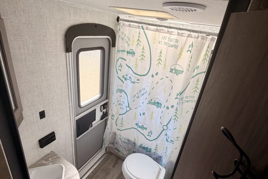 Compact RV rental bathroom featuring a functional toilet, sink, and a shower curtain: "Life is Better at the Campsite."