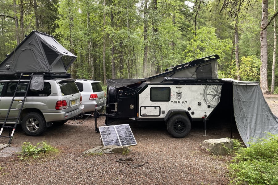 Explore the wilderness! RV rental featuring an SUV with a rooftop tent, overland trailer, and solar power setup in a lush forest.