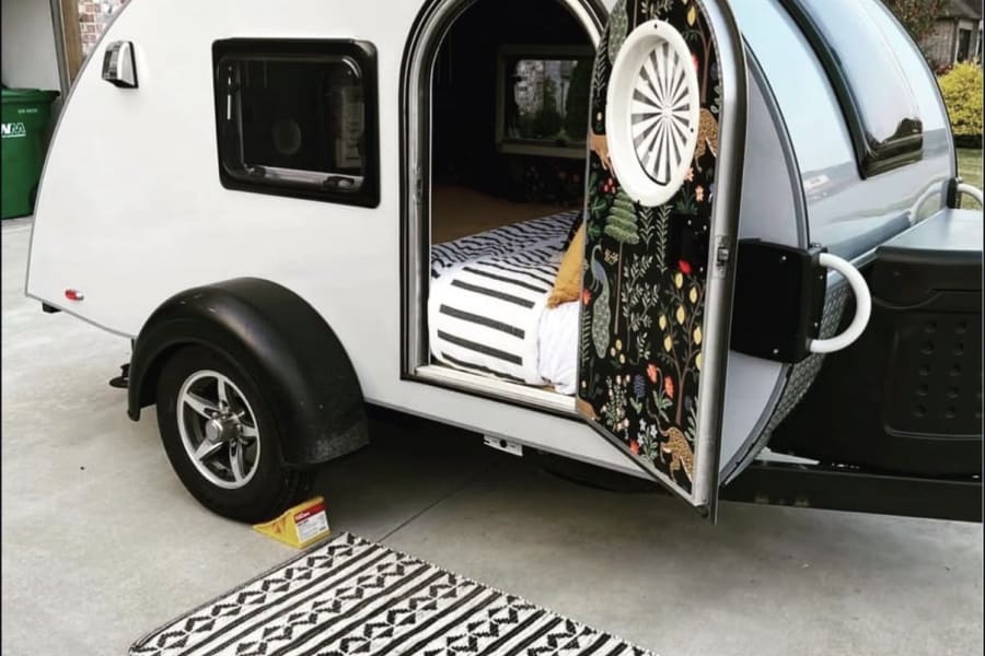 Stylish teardrop RV rental open, showing a bed & patterned door. Black & white rug out front, parked on a driveway.