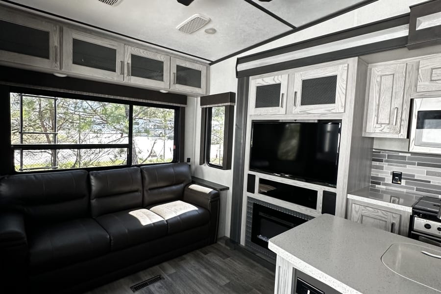Modern RV rental interior, featuring a spacious living area with a dark leather sofa, TV, fireplace, and a functional kitchen.