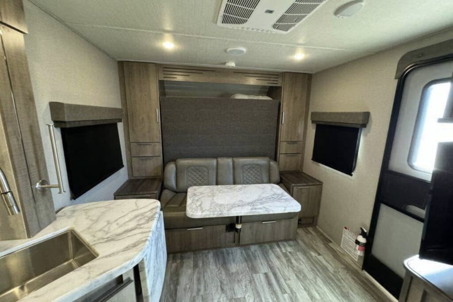 RV rental interior featuring a compact kitchen, dining booth, and wall-mounted bed. Wood-look floors, cabinetry, and small windows.