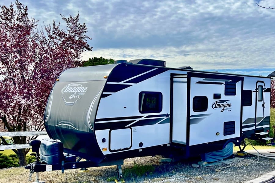 A modern Imagine XLS travel trailer, white with black accents, parked with its door open. Perfect for an rv rental, it also has a 'For Sale' sign.