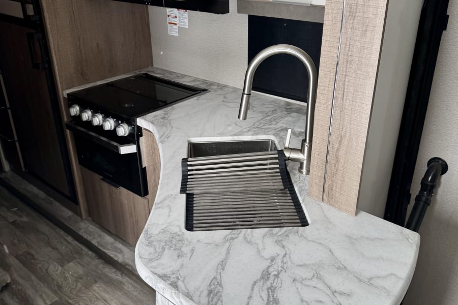 Compact RV rental kitchen featuring a microwave, stove, sink with drying rack, and marble-look countertop, ideal for travel.