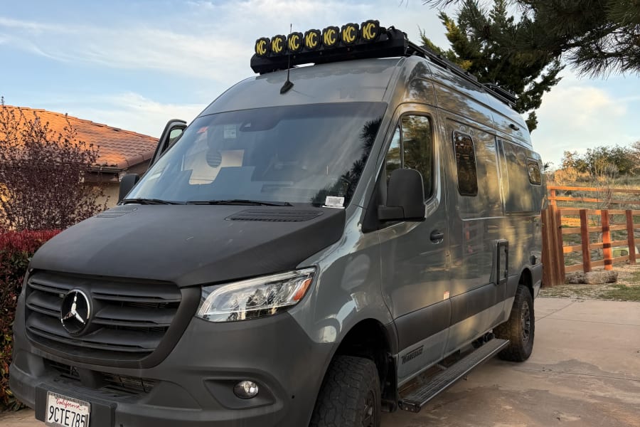 Adventure-ready gray Mercedes Sprinter rv rental, customized for off-road trips with a roof light bar & chunky tires, parked on a driveway.