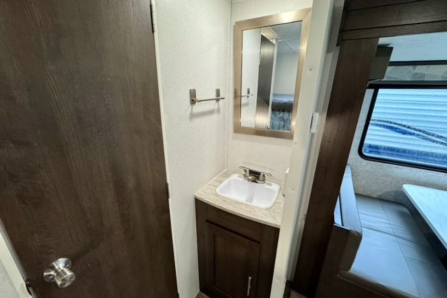 Compact RV rental bathroom with a sink, mirror, and towel bar, adjacent to a door and a dinette with a window.