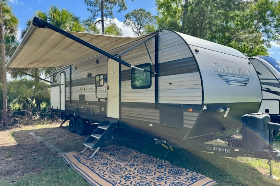 This Forest River Salem travel trailer, ideal for an RV rental, is set up with an extended awning and outdoor rug in a natural setting.