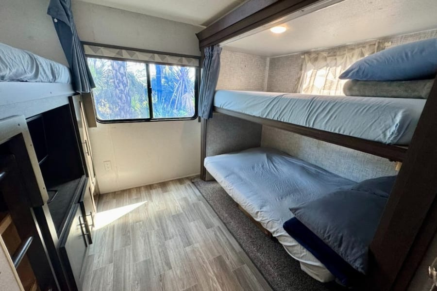 RV rental's bunk beds: Three cozy berths with light blue bedding, a window with outdoor views, and wood-look flooring.