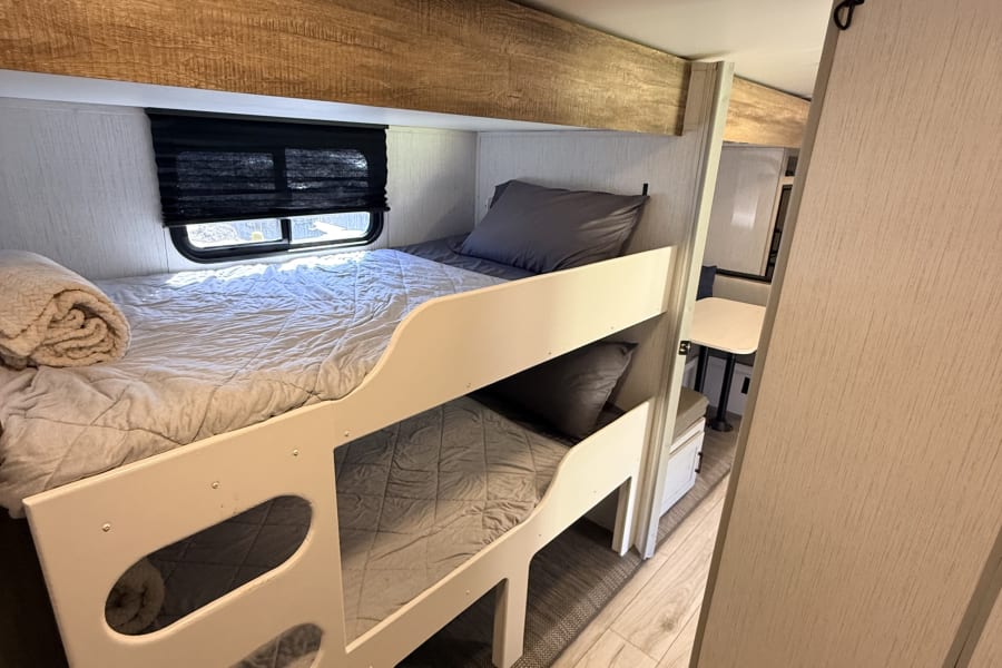 This RV rental offers cozy bunk beds with light gray bedding, a window with a blind, and wood accents for travel.