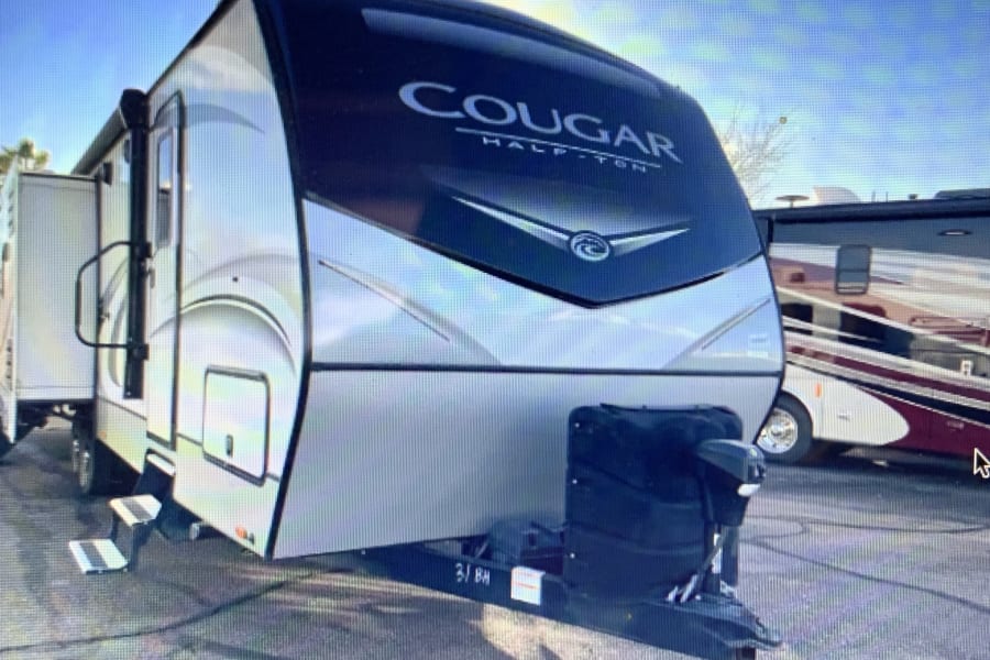 This Cougar Half-Ton travel trailer, perfect for an RV rental, is ready with its steps down and hitch, parked in sunlight.
