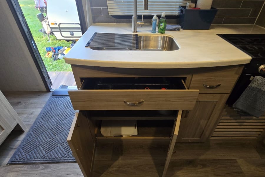 RV rental kitchen: sink with drying rack, open drawer/cabinet storage, and a view of the campsite outside.