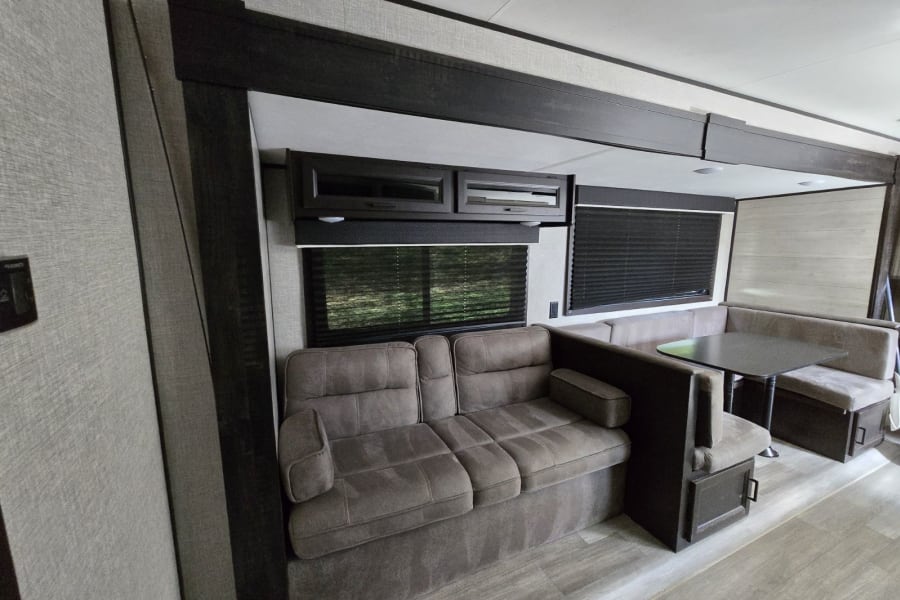 RV rental interior: A spacious living area features a comfortable sofa, dinette, overhead cabinets, and windows with blinds.