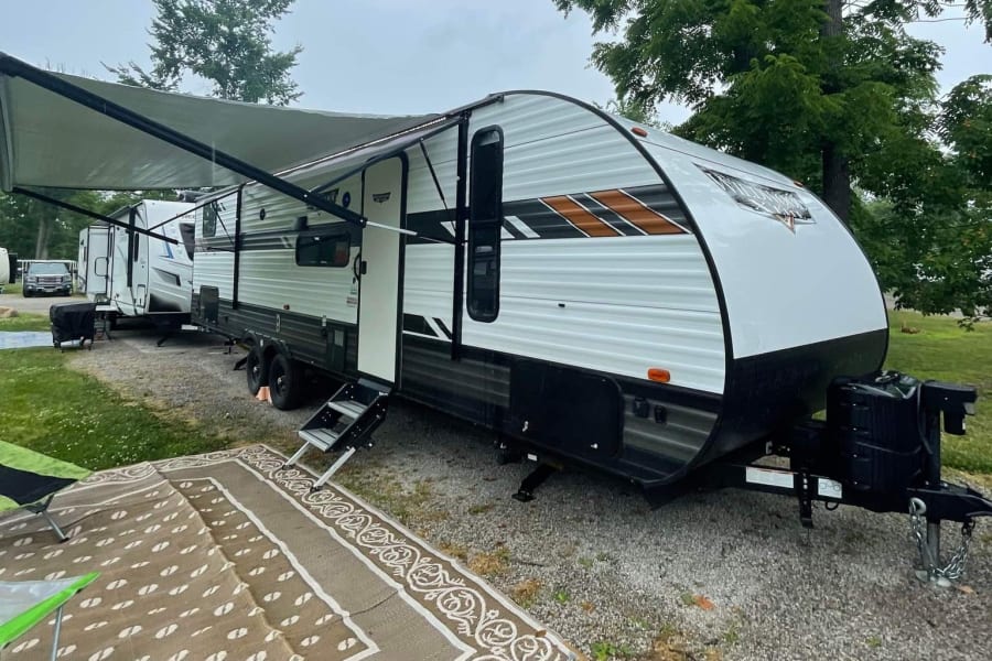 This white travel trailer RV rental, with an extended awning and outdoor rug, is set up on a gravel campsite for camping fun.