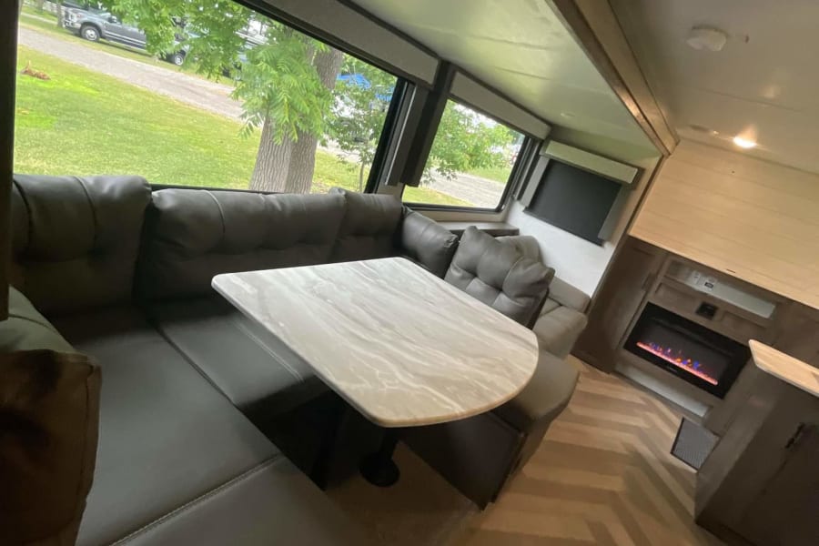 Modern rv rental: gray U-shaped dinette, marble-look table, large windows, electric fireplace, and herringbone floor.