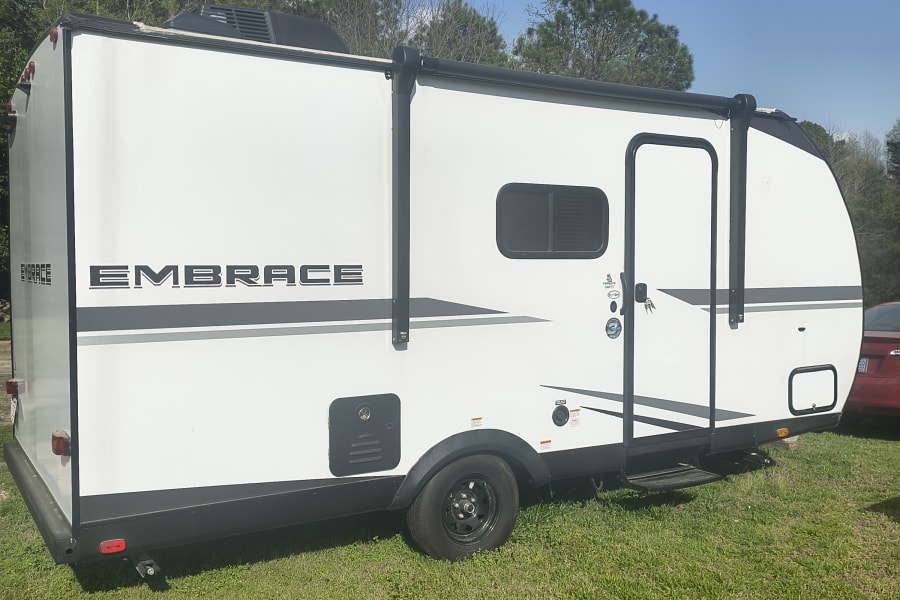 A white "EMBRACE" travel trailer with a black awning and door, parked on green grass. Ideal for an RV rental getaway.