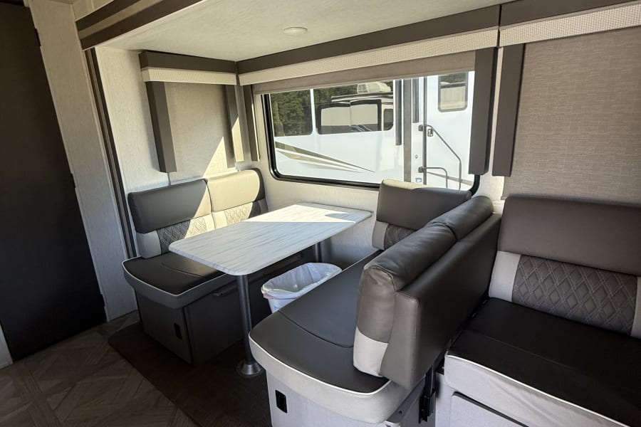 RV rental dining area with modern gray and white cushioned seats, a table, and window showing another camper outside.