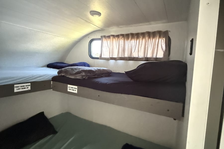 Inside an RV rental: a compact sleeping area with multiple bunk beds, a window with curtains, and overhead lighting.