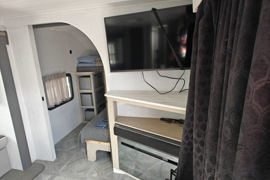 Modern RV rental interior: living area with TV, decorative fireplace, and a peek into the bedroom. Bright, compact design.