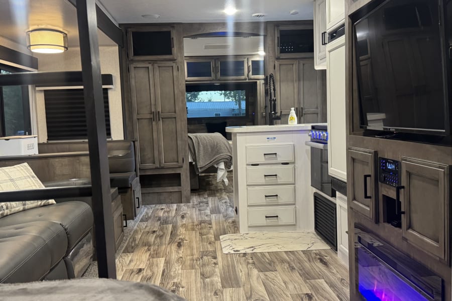 This modern RV rental interior showcases a kitchen island, entertainment unit with a fireplace, dinette, and wood-look floors.