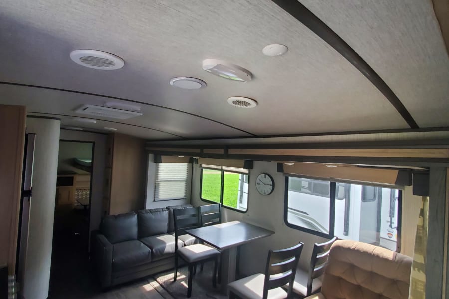 Spacious RV rental living area with a gray sofa, dining table, chairs, and a light brown recliner, ready for travel.