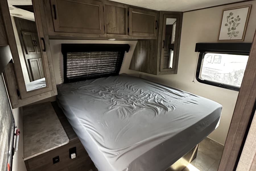 This RV rental bedroom features a grey bed, overhead wooden cabinets, a side window, and a built-in nightstand.