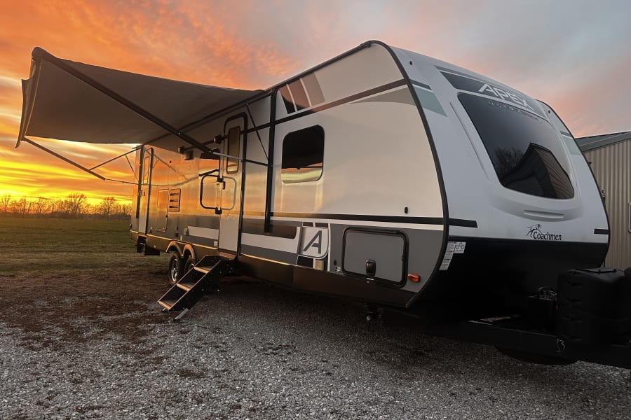 RV rental travel trailer with awning extended, glowing under a vibrant orange and yellow sunset by a field.