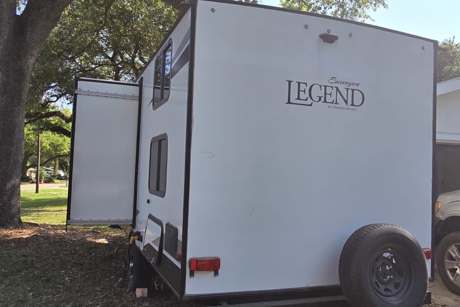 White Legend travel trailer, a spacious RV rental, parked under a sunny, leafy tree, perfect for outdoor camping.