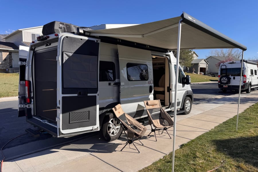 A white camper van RV rental, with its awning extended & chairs set up, creates a cozy outdoor spot on a sunny day.