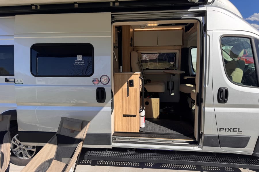 Explore this silver camper van rv rental! Side door open, showcasing a modern interior, extended awning, and outdoor chairs.