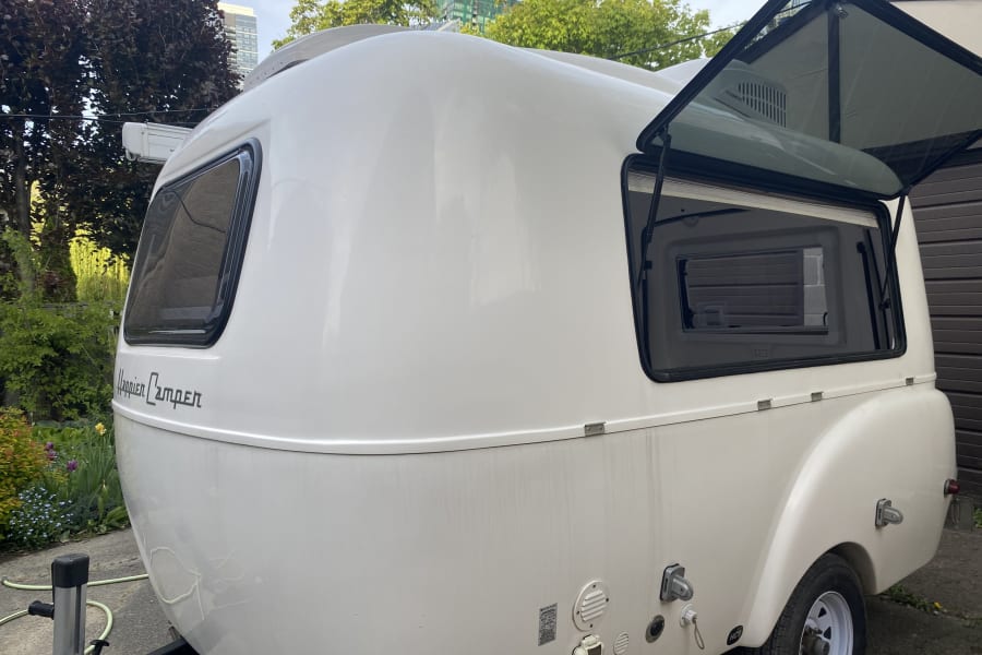 White Happier Camper RV rental trailer, showcasing its compact design, open window, and utility hookups.