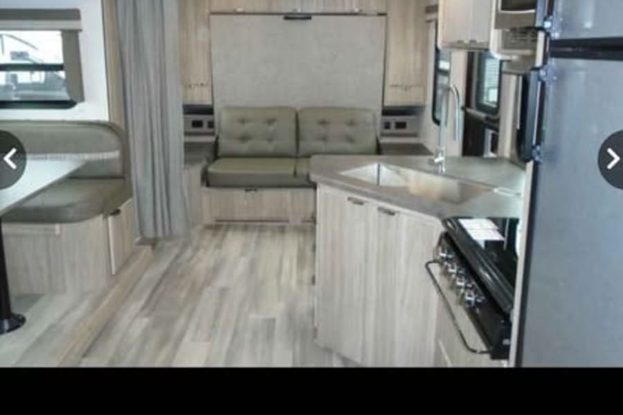 Modern RV rental interior with a compact kitchen, dinette, and sofa/fold-down bed in light wood and grey tones.