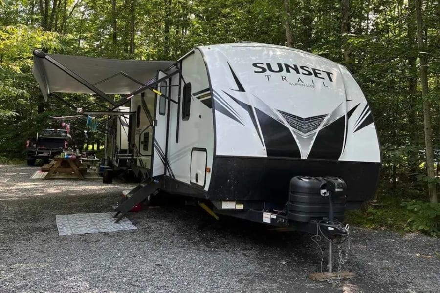 This Sunset Trail travel trailer RV rental is set up at a wooded campsite with an extended awning and picnic area.